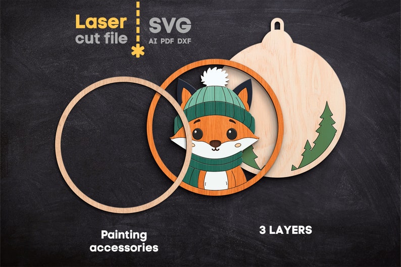 May include: A laser cut file SVG design for a three-layer wooden ornament featuring a fox wearing a teal beanie and scarf. The design includes a circular frame, a fox cutout, and a back piece with pine tree silhouettes. Text reads Laser cut file, SVG, AI, PDF, DXF, 3 LAYERS, and Painting accessories.