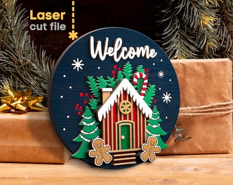 May include: A blue wooden round sign with the word "Welcome" in white lettering. The sign features a gingerbread house with red and white stripes, a green door, and a candy cane. Two gingerbread people stand on either side of the house. The sign is decorated with green trees, red berries, and white snowflakes.