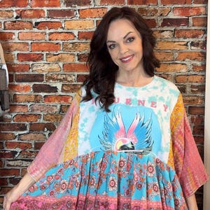 May include: A multi-colored patchwork tunic featuring a central graphic with the text JOURNEY and a winged design. The garment combines tie-dye patterns, floral prints, and various fabric textures in shades of blue, pink, and yellow.