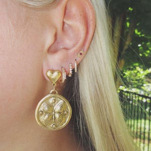 May include: A close-up of an ear adorned with multiple gold earrings. The earrings include a heart-shaped stud, a small hoop with clear stones, a small hoop with clear stones, a small hoop with clear stones, and a large gold pendant with a butterfly design.