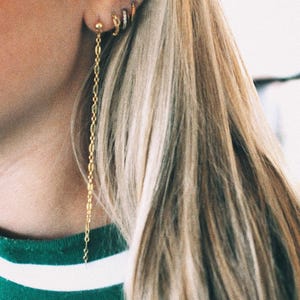 May include: Close-up of a person's ear adorned with multiple gold earrings. The ear has a small stud, a hoop with small stones, and a long chain earring. The person is wearing a green shirt with a white stripe.