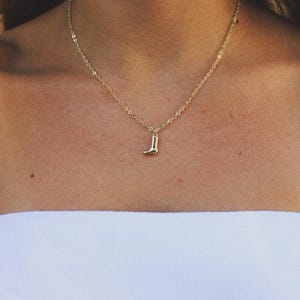 Puede incluir: Un collar dorado con un pequeño colgante de bota de vaquero. El colgante es una miniatura detallada de una bota de vaquero. El collar se lleva sobre la piel de una persona y un top blanco sin tirantes.