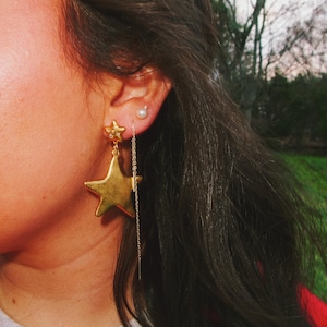 Könnte beinhalten: Goldene Sternohrringe mit einer herabhängenden Kette. Ein Ohrring hat einen kleinen Sternstecker, der andere einen großen, fünfzackigen Sternanhänger. Die Ohrringe werden von einer Person mit dunklem Haar und einem roten Oberteil getragen.