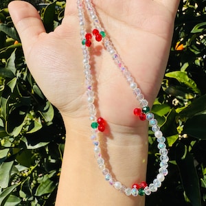 Puede incluir: Un charm de teléfono de cuentas blancas con cuentas rojas y verdes con forma de cerezas.