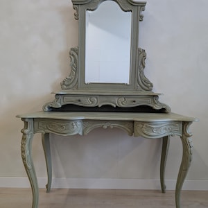 May include: A sage green vanity table featuring a tall, arched mirror with ornate carved detailing. The piece includes a wide tabletop, two small drawers beneath the mirror, and two additional drawers on the main desk, all supported by four curved legs.