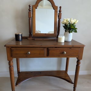 May include: A wooden vanity table with a medium brown finish, featuring two drawers with round knobs, four turned legs, and a lower shelf. A matching arched swivel mirror sits on top, accompanied by a black candle, a white candle, and a vase of white roses.