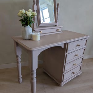 May include: A mauve-coloured vanity desk featuring a rectangular swivel mirror, a small drawer with two gold-toned knobs, and a side cabinet with four stacked drawers. The desk has turned legs and is topped with a white vase of roses and a glass candle.