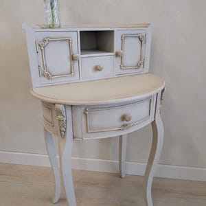 May include: A pale grey, antique-style side table with a curved front and cabriole legs. The table features a small cabinet with two doors and a drawer, all adorned with gold-coloured trim and knobs. A vase with a pink flower sits on top.