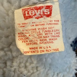 Puede incluir: Primer plano de una etiqueta de Levi's con texto y logotipo rojos sobre fondo blanco. La etiqueta incluye instrucciones de lavado y el texto "MADE IN U.S.A.". La etiqueta est&aacute; adherida a una tela azul claro y difusa.