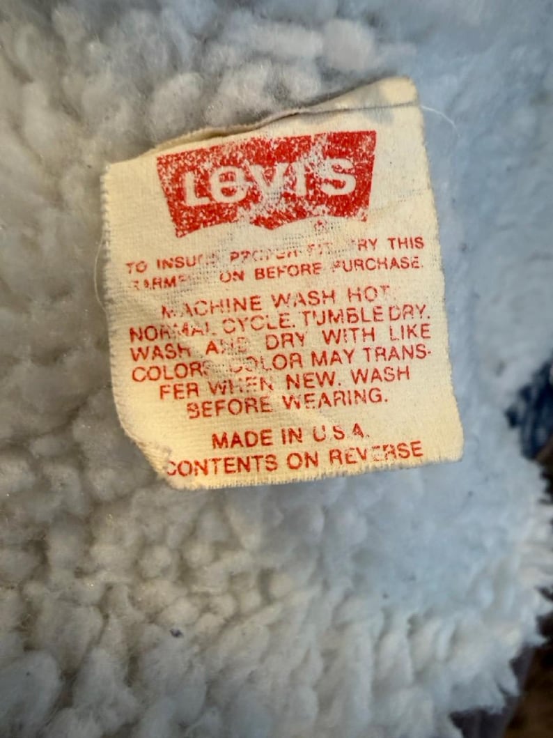 Puede incluir: Primer plano de una etiqueta de Levi's con texto y logotipo rojos sobre fondo blanco. La etiqueta incluye instrucciones de lavado y el texto "MADE IN U.S.A.". La etiqueta est&aacute; adherida a una tela azul claro y difusa.