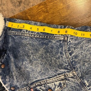 Puede incluir: Chaqueta vaquera con lavado azul claro y forro de borreguillo blanco. La chaqueta tiene dos bolsillos en el pecho y una etiqueta roja. El hombro mide aproximadamente 23 cm.