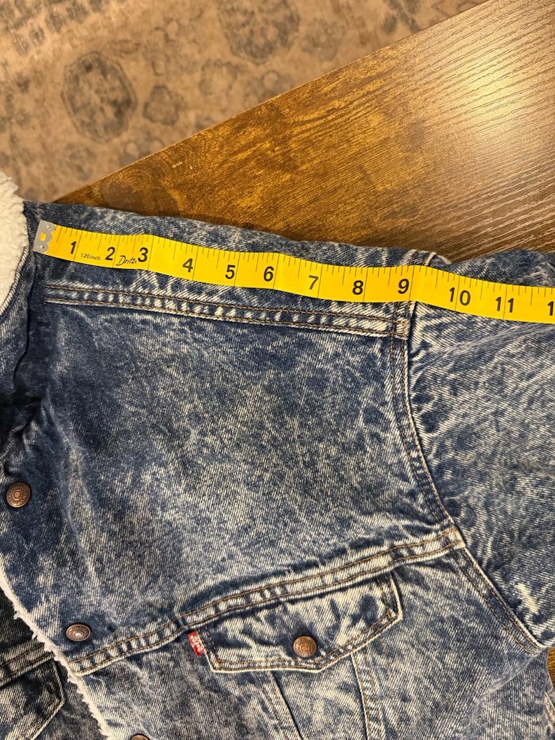 Puede incluir: Chaqueta vaquera con lavado azul claro y forro de borreguillo blanco. La chaqueta tiene dos bolsillos en el pecho y una etiqueta roja. El hombro mide aproximadamente 23 cm.