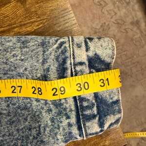 Puede incluir: Primer plano de una manga de chaqueta vaquera con una cinta m&eacute;trica amarilla que muestra una longitud de aproximadamente 76 cm. La chaqueta tiene un estampado de lavado &aacute;cido azul y blanco y est&aacute; sobre una superficie de madera.