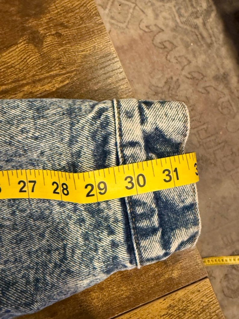 Puede incluir: Primer plano de una manga de chaqueta vaquera con una cinta m&eacute;trica amarilla que muestra una longitud de aproximadamente 76 cm. La chaqueta tiene un estampado de lavado &aacute;cido azul y blanco y est&aacute; sobre una superficie de madera.