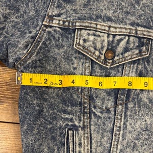 Puede incluir: Primer plano de una chaqueta vaquera azul con efecto lavado a la piedra. La chaqueta tiene un bolsillo en el pecho con un bot&oacute;n y una cinta m&eacute;trica amarilla que muestra una longitud de aproximadamente 23 cm.