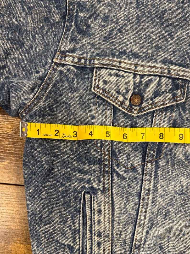 Puede incluir: Primer plano de una chaqueta vaquera azul con efecto lavado a la piedra. La chaqueta tiene un bolsillo en el pecho con un bot&oacute;n y una cinta m&eacute;trica amarilla que muestra una longitud de aproximadamente 23 cm.