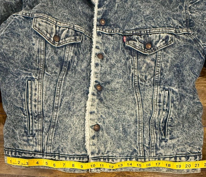 Puede incluir: Chaqueta vaquera con lavado &aacute;cido y forro de borreguillo color crema. La chaqueta tiene dos bolsillos en el pecho y dos bolsillos laterales. La chaqueta est&aacute; abotonada y tiene una cinta m&eacute;trica amarilla en la parte inferior. La chaqueta mide unos 53 cm en la parte inferior.