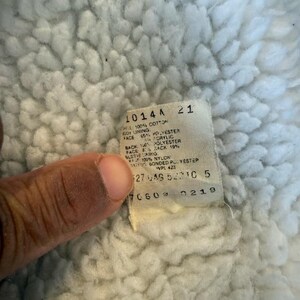 Puede incluir: Primer plano de una tela blanca texturizada con una etiqueta adjunta. La etiqueta tiene texto que incluye "1014A" y detalles de la composici&oacute;n de la tela como "100% Algod&oacute;n" y "65% Poli&eacute;ster". La tela parece ser un forro.