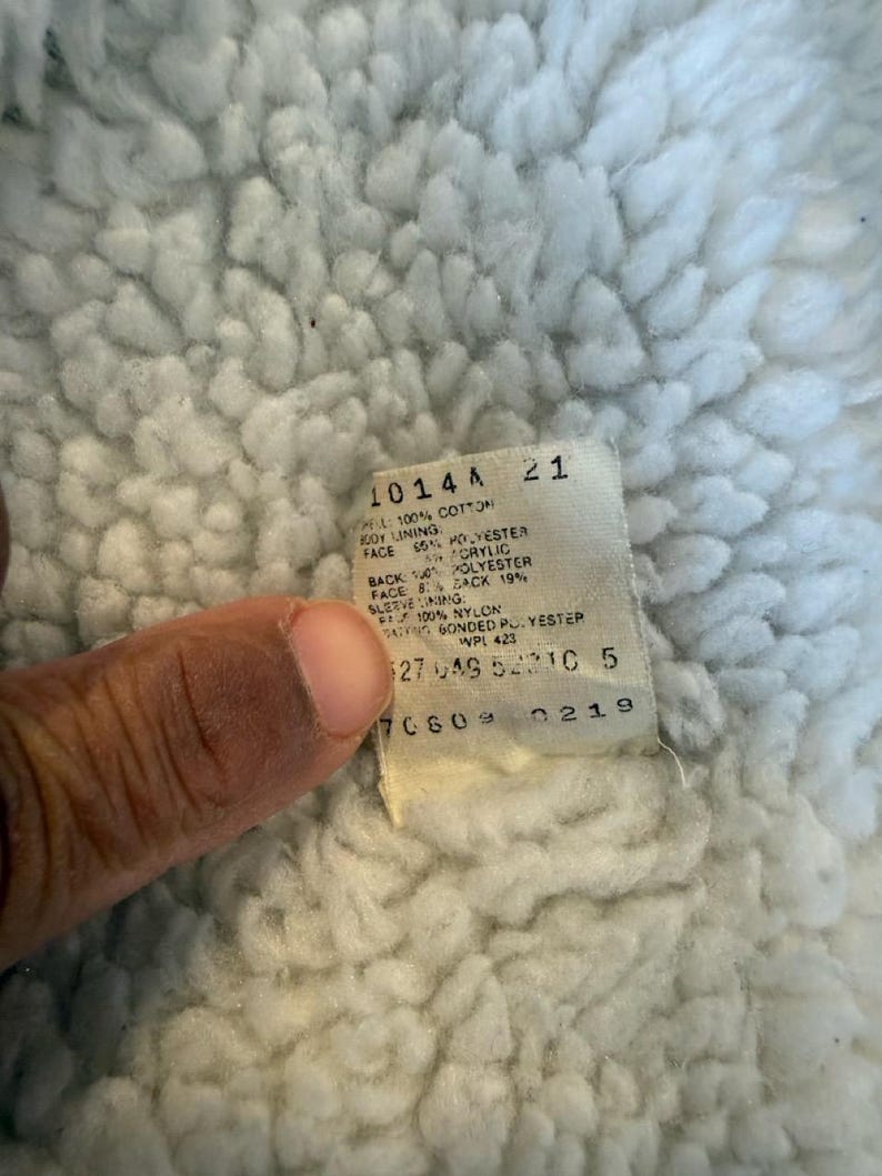 Puede incluir: Primer plano de una tela blanca texturizada con una etiqueta adjunta. La etiqueta tiene texto que incluye "1014A" y detalles de la composici&oacute;n de la tela como "100% Algod&oacute;n" y "65% Poli&eacute;ster". La tela parece ser un forro.