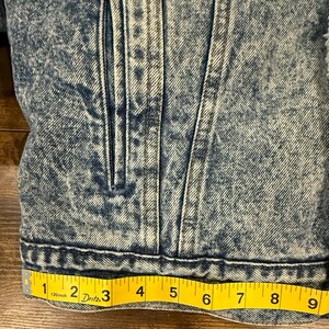 Puede incluir: Primer plano de una chaqueta vaquera azul con efecto lavado a la piedra. La chaqueta tiene cierre de botones, bolsillos y una cinta m&eacute;trica amarilla que muestra una longitud de 23 cm.