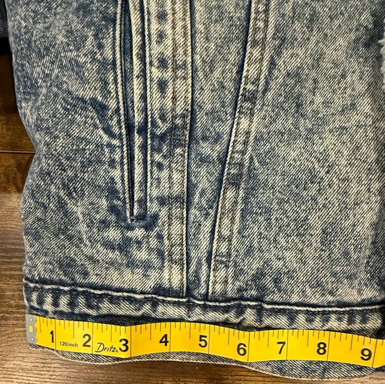 Puede incluir: Primer plano de una chaqueta vaquera azul con efecto lavado a la piedra. La chaqueta tiene cierre de botones, bolsillos y una cinta m&eacute;trica amarilla que muestra una longitud de 23 cm.