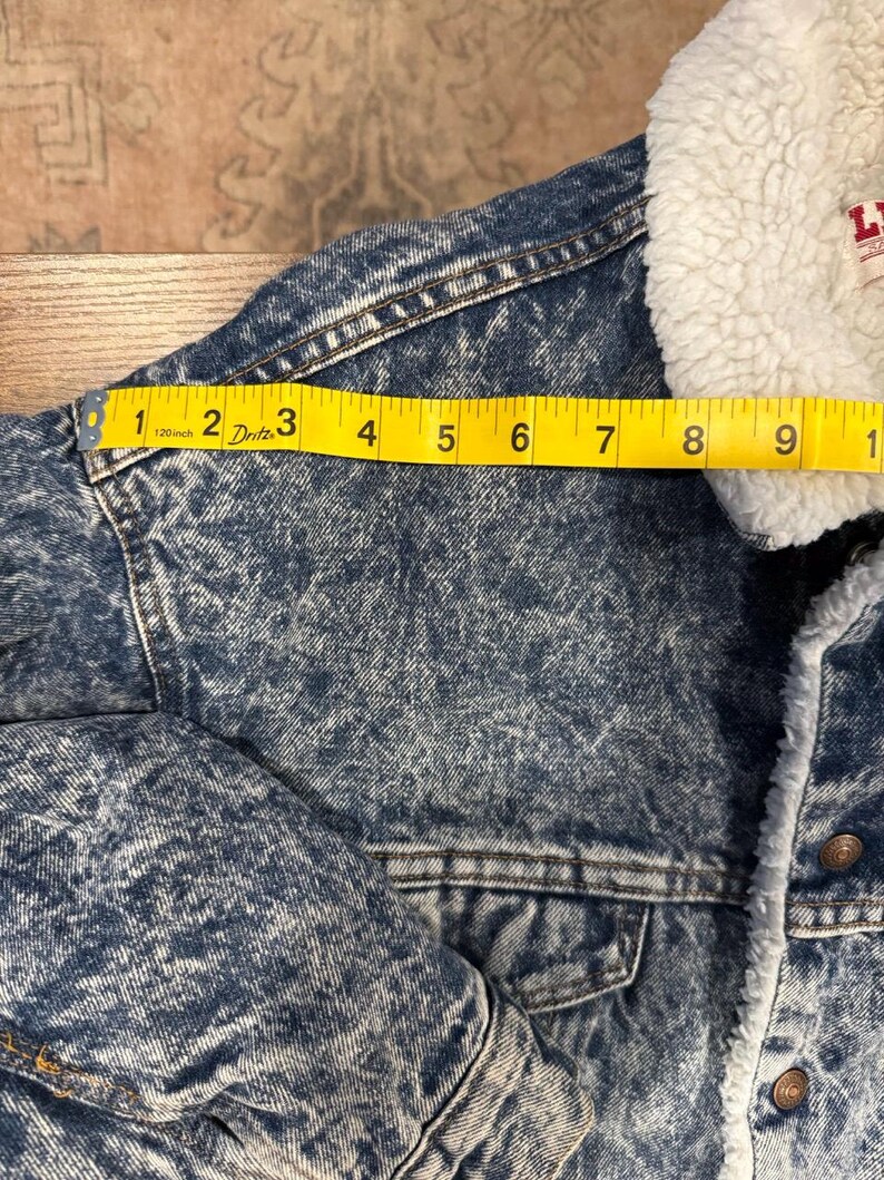 Puede incluir: Chaqueta vaquera con cuello de borreguillo blanco. La chaqueta est&aacute; lavada a la piedra y tiene una cinta m&eacute;trica amarilla sobre el hombro, que muestra una medida de aproximadamente 23 cm.