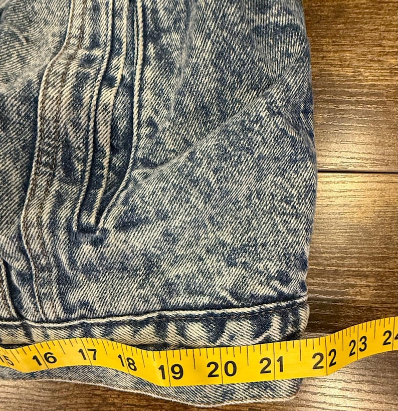Puede incluir: Primer plano de una chaqueta vaquera azul con un ligero lavado &aacute;cido. Una cinta m&eacute;trica amarilla muestra una longitud de aproximadamente 40 a 61 cm. La chaqueta tiene bolsillos y costuras.