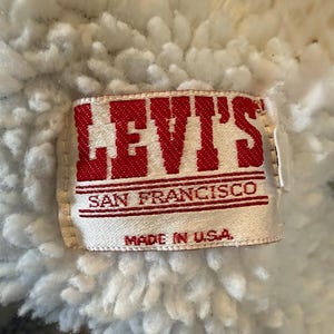 Puede incluir: Primer plano de una etiqueta de Levi's, con el nombre de la marca en letras rojas. La etiqueta dice "SAN FRANCISCO" y "MADE IN U.S.A." sobre un fondo blanco. La etiqueta est&aacute; colocada sobre un fondo de tela blanca y esponjosa.
