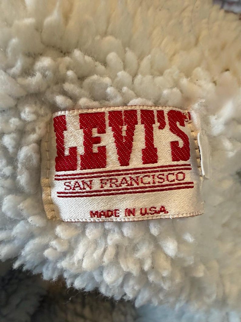 Puede incluir: Primer plano de una etiqueta de Levi's, con el nombre de la marca en letras rojas. La etiqueta dice "SAN FRANCISCO" y "MADE IN U.S.A." sobre un fondo blanco. La etiqueta est&aacute; colocada sobre un fondo de tela blanca y esponjosa.