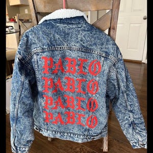 Puede incluir: Chaqueta vaquera con lavado &aacute;cido y cuello de borreguillo blanco. La parte trasera de la chaqueta presenta el nombre "PABLO" en letras rojas grandes. La chaqueta est&aacute; colgada en una silla de madera.