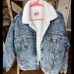 Puede incluir: Chaqueta vaquera azul con forro y cuello de borreguillo blanco. La chaqueta tiene cierre de botones, dos bolsillos en el pecho y dos bolsillos laterales. El logo de Levi's es visible.