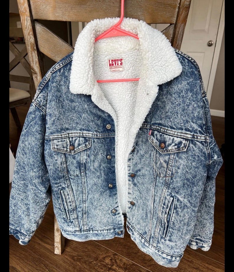 Puede incluir: Chaqueta vaquera azul con forro y cuello de borreguillo blanco. La chaqueta tiene cierre de botones, dos bolsillos en el pecho y dos bolsillos laterales. El logo de Levi's es visible.