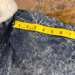 Puede incluir: Primer plano de una chaqueta vaquera con un cuello blanco y esponjoso. La chaqueta es azul lavado a la piedra con una cinta m&eacute;trica amarilla que muestra una medida de aproximadamente 18 cm.