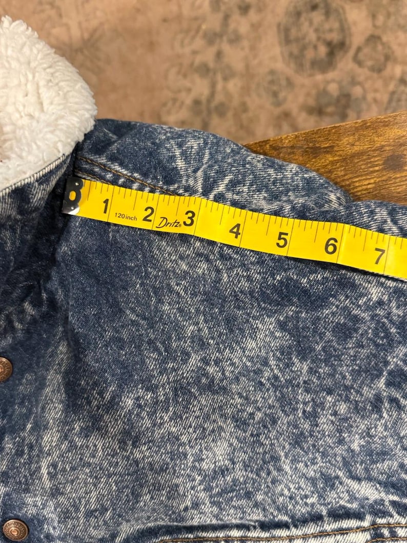 Puede incluir: Primer plano de una chaqueta vaquera con un cuello blanco y esponjoso. La chaqueta es azul lavado a la piedra con una cinta m&eacute;trica amarilla que muestra una medida de aproximadamente 18 cm.