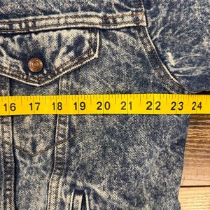 Puede incluir: Primer plano de una chaqueta vaquera azul con un bolsillo abotonado y un bot&oacute;n met&aacute;lico. La chaqueta tiene un aspecto descolorido, lavado a la piedra. Una cinta m&eacute;trica amarilla se coloca sobre la chaqueta, mostrando medidas de 40,6 a 61 cm.