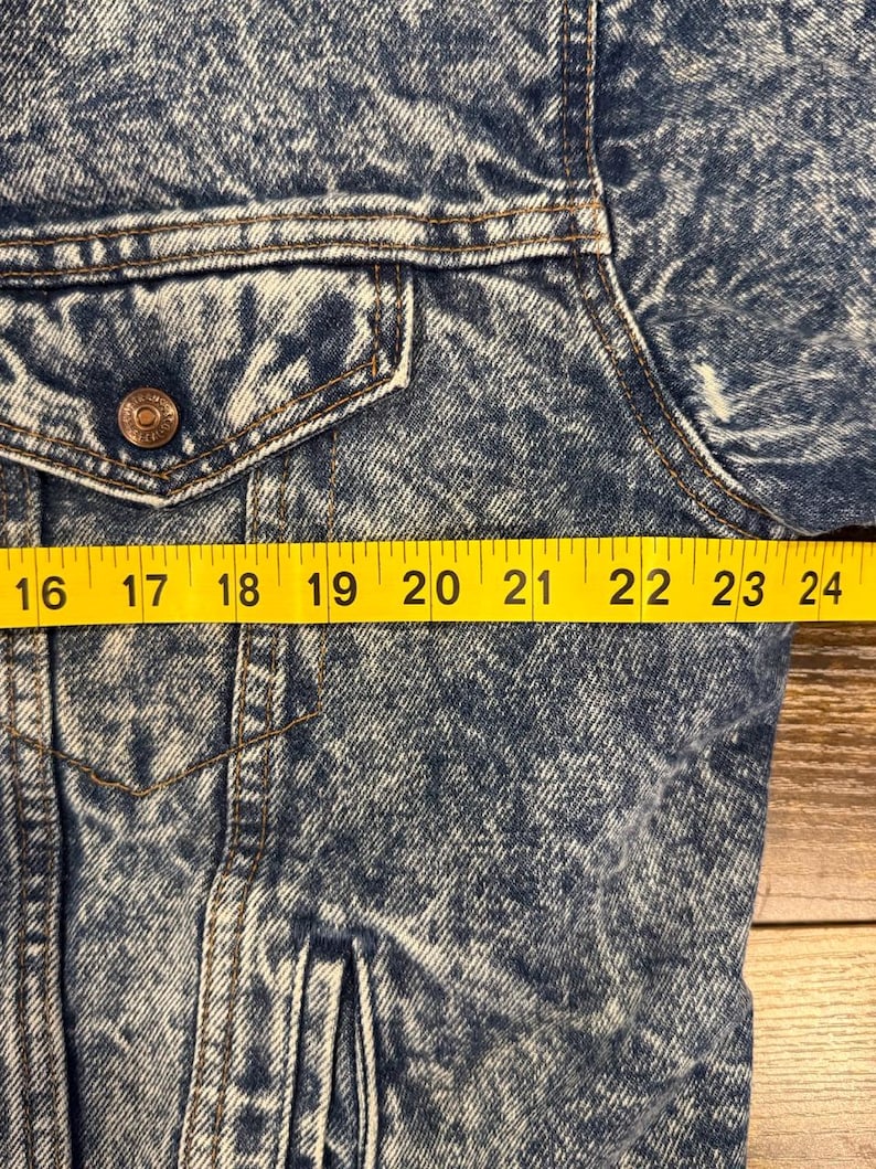 Puede incluir: Primer plano de una chaqueta vaquera azul con un bolsillo abotonado y un bot&oacute;n met&aacute;lico. La chaqueta tiene un aspecto descolorido, lavado a la piedra. Una cinta m&eacute;trica amarilla se coloca sobre la chaqueta, mostrando medidas de 40,6 a 61 cm.