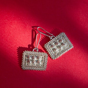 May include: A pair of silver-toned rectangular earrings with intricate filigree detailing and small red accents. The earrings have a hook closure and are displayed against a vibrant red background, showcasing their ornate design.