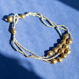 Op de afbeelding: Een gouden en zilveren kralenarmband met een geometrisch ontwerp. De armband heeft meerdere strengen kleine zilveren kralen en grotere goudkleurige, facetgeslepen kralen. De armband wordt gepresenteerd op een blauw oppervlak.