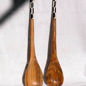 May include: A pair of wooden salad servers. The spoons have a natural wood finish with a giraffe-like pattern on the handles. One spoon is solid, and the other has slots for draining.