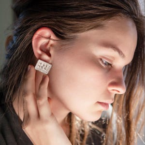 May include: A close-up of a person wearing a silver square-shaped earring with a textured design.