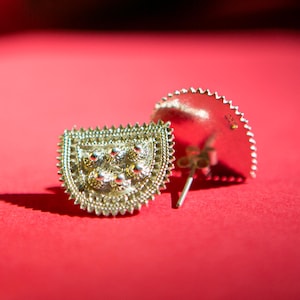 May include: A pair of silver earrings with a geometric design. The earrings are shaped like a heart and have a textured surface. The earrings are on a red background.