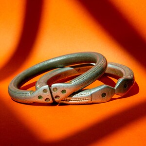 Peut inclure: Deux bracelets articulés argentés avec des motifs décoratifs de points et de lignes. Les bracelets sont posés sur un fond orange vif, créant un contraste saisissant. Le métal semble vieilli, leur donnant un aspect vintage. Chaque bracelet mesure environ 6 cm de diamètre.