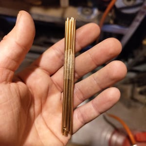 May include: A collection of slender, gold-coloured metal rods, secured with clear tape. The rods are cylindrical and likely made of brass or a similar alloy. The background is blurred, suggesting an indoor setting.