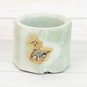 Puede incluir: Un pequeño macetero de hormigón de color verde claro con un diseño de pato en hoja de oro. El macetero tiene una forma hexagonal y es perfecto para plantas pequeñas o suculentas.