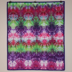 May include: A tie-dye quilt with a vibrant pattern of pink, purple, and green hues. The quilt features a repeating floral-like design, bordered by a deep purple edge. The quilt is hung on a wall with clips.