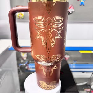 May include: A brown travel mug with a handle and a black lid. The mug is decorated with gold-colored geometric patterns. The handle and the rim of the mug are also brown. The mug is sitting on a white platform.
