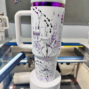 May include: A white travel mug with a purple lid and a metallic purple band around the top. The mug has a black and white design featuring the Hogwarts castle, the Deathly Hallows symbol, and the number 9 3/4.