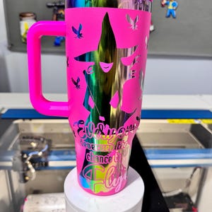 May include: A pink stainless steel tumbler with a handle and a straw. The tumbler features a design of a witch with a star and butterflies. The text on the tumbler reads "Everyone deserves the chance to fly".