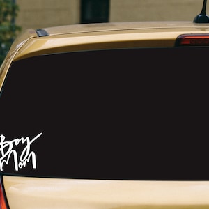May include: White vinyl decal reading "Boy Mom" in a cursive font, applied to the rear window of a gold car. The decal is placed on the lower left of the window, against a black background.