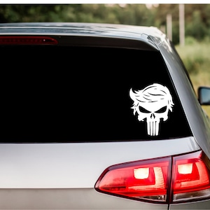 May include: A white vinyl decal of a skull with a hairstyle resembling that of Donald Trump. The decal is applied to the rear window of a silver car.